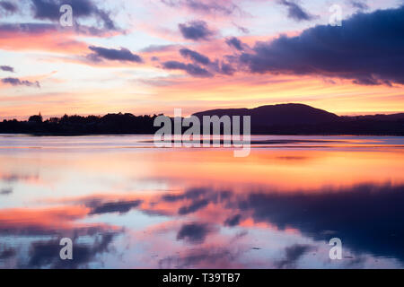 Coucher du soleil brillant avec des couleurs terre lointaine silhouette et image miroir reflète dans la baie calme de l'eau longue exposition. Banque D'Images