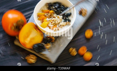 Motion Blur en arrière-plan de l'extrait d'avoine avec kaki, pruneaux, abricots secs, noix et graines de sésame sur une table en bois noir. Banque D'Images