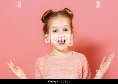 Closeup portrait of attractive cheerful charmante petite fille aux brioches sur fond rose. L'enfant de surprise et répandre les deux mains sur les côtés. Banque D'Images