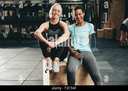 Deux jeunes femmes dans les vêtements de sport tout en restant assis souriant ensemble dans une salle de sport après l'entraînement Banque D'Images