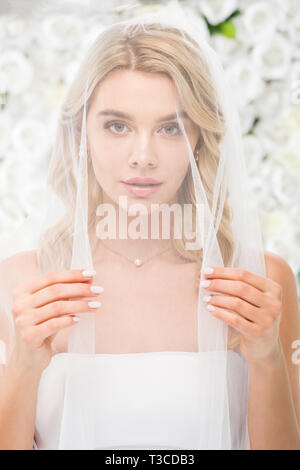 Belle jeune femme à visage couvert avec voile transparent looking at camera on white floral background Banque D'Images