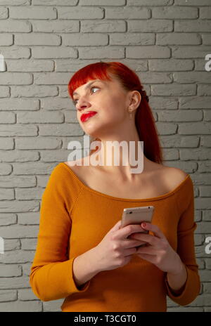 Jeune fille rousse est en train de lire quelque chose de façon réfléchie sur son smartphone contre un mur de brique gris Banque D'Images