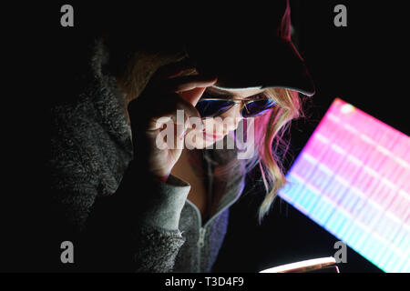 Fille Hacker dans un chapeau et des lunettes de soleil devant un ordinateur en neon light tient en main du smartphone Banque D'Images