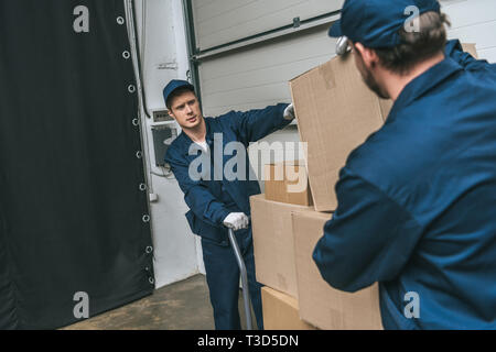 Deux movers en uniforme à l'aide de camion de main durant le transport de boîtes de carton dans l'entrepôt avec copie espace Banque D'Images