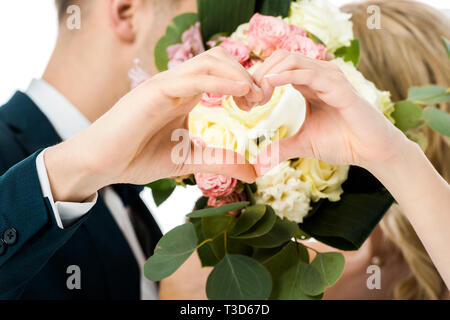Focus sélectif du marié et mariée montrant coeur signe avec les mains, tout en masquant les visages derrière mariage bouquet isolated on white Banque D'Images