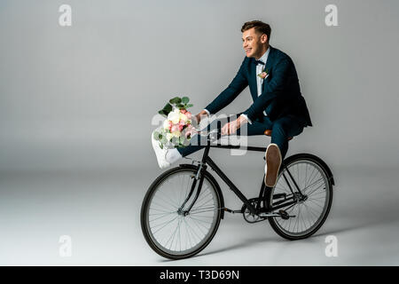 Happy groom dans un élégant costume et sneakers riding bike et la tenue mariage bouquet sur fond gris Banque D'Images