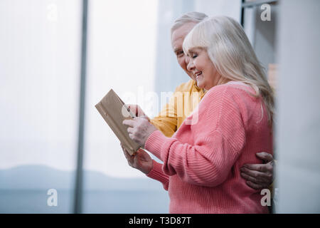 Happy senior couple dans des vêtements colorés reading book at home with copy space Banque D'Images