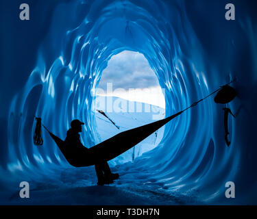 Silhouette d'un homme assis dans un hamac à l'intérieur d'une grotte de glace en Alaska. Son casque, piolets, cordes et accrocher au murs bleu profond de la grotte sur le Banque D'Images