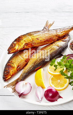 Ensemble de harengs fumés à chaud avec les tranches de citron, le persil et l'oignon rouge sur une assiette blanche sur une table en bois, vue verticale de ci-dessus, close-up Banque D'Images