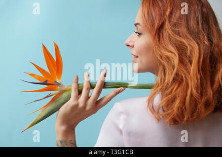 Redhead happy girl holding sur son épaule une fleur orangée strelitzia autour d'un fond bleu avec l'espace pour le texte. Mise en page pour vos idées créatives. Banque D'Images