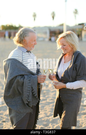 Deux pensionnés debout avec verres de champagne sur la plage de sable et le port de tissus écossais. Concept de se reposer à l'air libre et couple de personnes âgées sur pique-nique. Banque D'Images