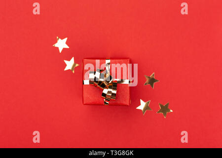 Boîte cadeau rouge avec archet d'or sur fond rouge décoré de confettis Etoiles d'or. Vue de dessus, de Noël et de vacances de style minimal concept. Banque D'Images