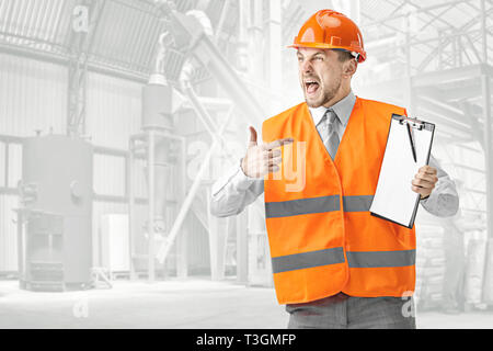 Le constructeur en colère dans un gilet et casque de construction d'orange de crier contre l'arrière-plan industriel. Le spécialiste de la sécurité, l'ingénieur, l'industrie, l'architecture, le manager, l'occupation, l'homme d'affaires, concept d'emploi Banque D'Images