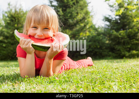 Portrait of elementary age girl enjoying eating tranche de pastèque juteuse allongé sur un pré, dans un jardin d'été Banque D'Images