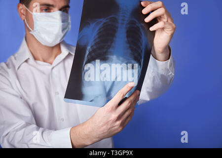 Médecin homme en blouse blanche et masque holding x-ray ou de roentgen, poumons, graphie image sur fond bleu Banque D'Images
