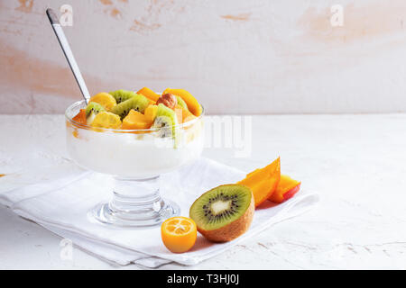 Bol en verre sur la tige avec du yogourt, des tranches de fruits : kumquat, kiwi, mangue et avec cuillère métallique est debout sur une serviette blanche sur tableau blanc. Copier l'espace. H Banque D'Images