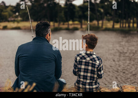 Vue arrière d'un homme assis avec son kid holding des cannes à pêche. Père et fils, assis sur le bord d'un lac de pêche de loisir de dépenser. Banque D'Images