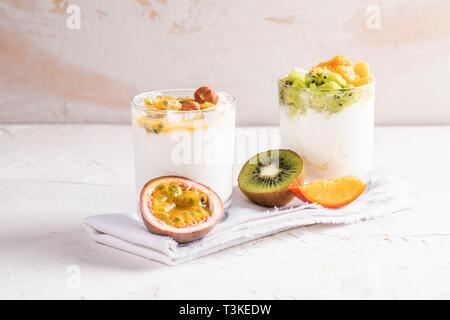 Deux verres de blanc des yaourts et des fruits frais : Purée de fruits de la passion, kiwi et mangue sont debout sur une serviette de cuisine blanc sur blanc bac texturé Banque D'Images