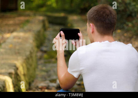 Jeune homme à prendre des photos à l'aide de son appareil photo téléphone intelligent dans la nature, parc et forêt, une petite rivière, un ruisseau. Homme de main holding smart phone. Banque D'Images