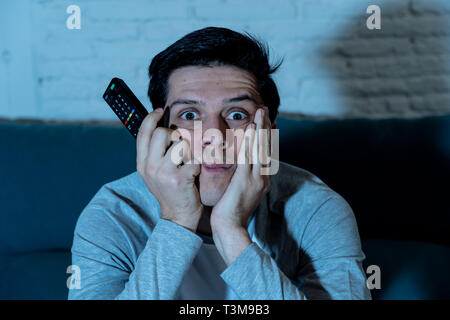 Jeune homme s'ennuie sur table à l'aide de la télécommande du téléviseur pour un autre film ou un zapping sport live tard dans la nuit. À boire sans sommeil et désintéressé b Banque D'Images