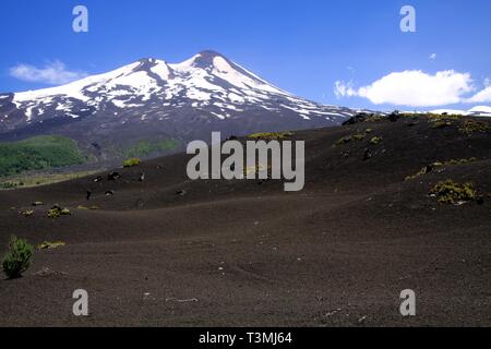 Large champ de lave volcanique sur le pic de cendres de volcan Llaima noir avec des taches et des bandes de neige et de glace contrastant avec ciel bleu - PN Conguillio Banque D'Images