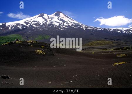 Large champ de lave volcanique sur le pic de cendres de volcan Llaima noir avec des taches et des bandes de neige et de glace contrastant avec ciel bleu - PN Conguillio Banque D'Images