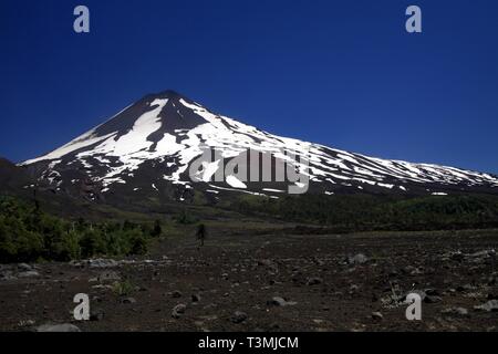 Large champ de lave volcanique sur le pic de cendres de volcan Llaima noir avec des taches et des bandes de neige et de glace contrastant avec ciel bleu - PN Conguillio Banque D'Images