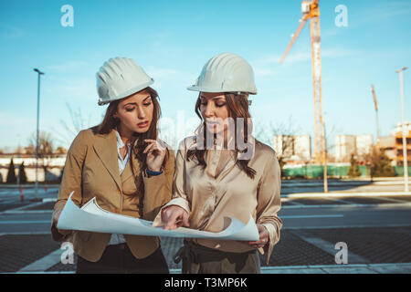 Les jeunes femmes ingénieurs réussie avec des casques blancs et bleus dans leurs mains sont l'analyse de nouveau projet au chantier de construction. Banque D'Images