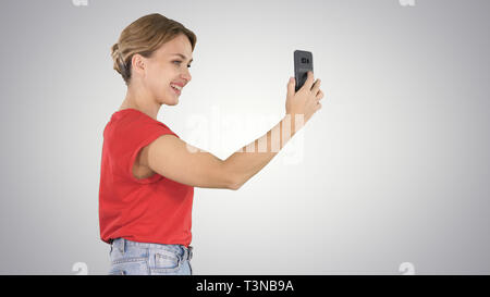 Belle jeune femme marche et tenant un smartphone jusqu'à prendre des photos et les autoportraits sur fond dégradé. Banque D'Images