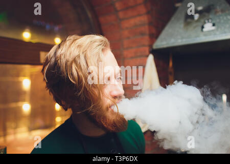 Le mec fume, exhale la fumée. La vapeur provenant d'une cigarette électronique. Portrait d'un jeune homme dans la fumée. Vortex vapeur coups à la main avec des taches de li Banque D'Images