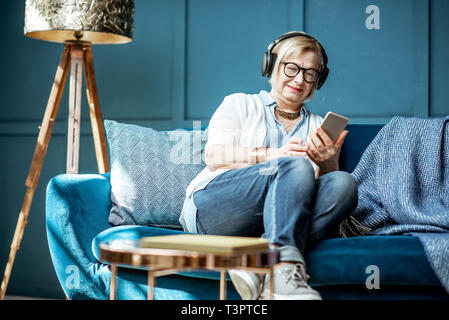 Hauts femme vêtue de désinvolture à écouter la musique au casque et le téléphone sur le canapé à la maison Banque D'Images