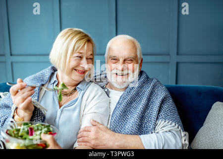 Joli couple manger salade saine en position assise recouverte d'un plaid sur le canapé à la maison Banque D'Images
