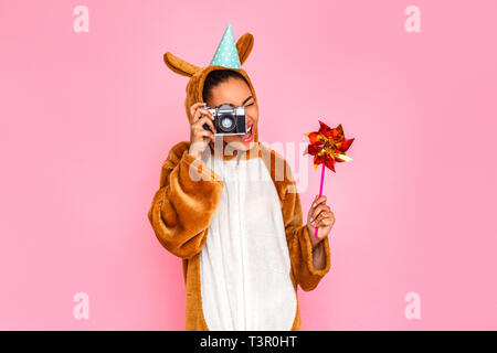 Jeune femme en bunny kigurumi wearing party hat standing isolé sur fond rose holding pinwheel prendre des photos sur l'appareil photo en souriant heureux Banque D'Images