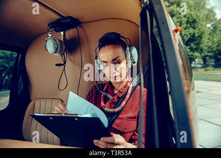 Dark-haired woman concentré de feuilleter des documents tout en portant des écouteurs Banque D'Images