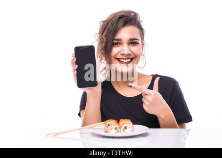 Smiling young asian woman eating sushi à la table isolated over white background, montrant écran blanc téléphone mobile Banque D'Images