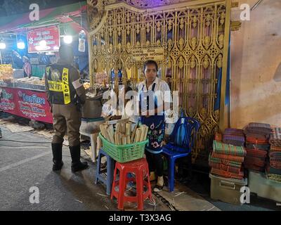 05 mars 2019, la Thaïlande, Karon Beach : un policier se tient à côté d'une femme vendant des desserts au marché du temple dans la plage de Karon. Le temple marché de plusieurs jours a toujours lieu autour de la fête bouddhiste Magha Puja. En ce jour, l'Eveillé est dit avoir livré un important premier sermon. Le marché de nuit est populaire auprès des touristes et des Thaïlandais. À côté de souvenirs et de faux produits il y a de nombreux stands de nourriture thaïe spécialités. Le marché du temple est situé dans la zone d'un temple à Karon. Une version plus petite du marché a lieu au même endroit l'ensemble de l'année comme un marché hebdomadaire. Banque D'Images