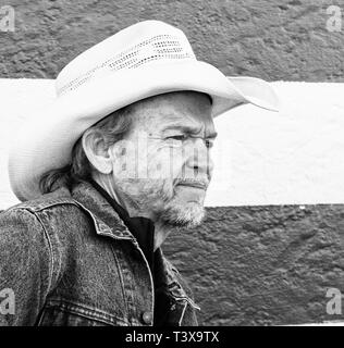 Old cowboy avec chapeau de cowboy et barbe grise a l'air sur le côté en se tenant debout à côté d'un mur à rayures blanches et noires. Banque D'Images