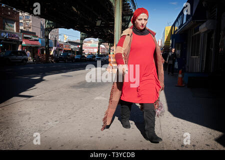 Une jolie femme en rouge portant un long manteau et de bottes hautes promenades au el sur Roosevelt Avenue à Jackson Heights, Queens, New York. Banque D'Images