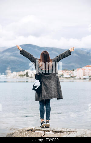 Une jeune fille se tient au large de la côte de la mer et regarde au loin et soulève ses mains montrant comment elle est heureuse. Banque D'Images