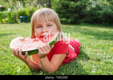 Portrait of elementary age girl enjoying eating tranche de pastèque juteuse allongé sur un pré, dans un jardin d'été Banque D'Images