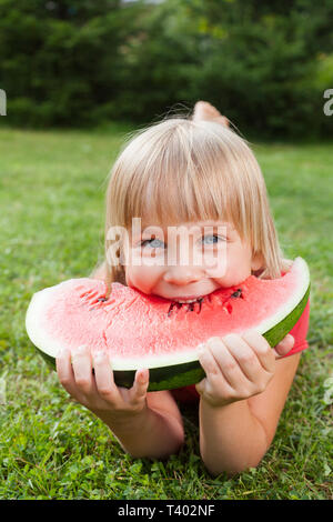 Portrait of elementary age girl enjoying eating tranche de pastèque juteuse allongé sur un pré, dans un jardin d'été Banque D'Images