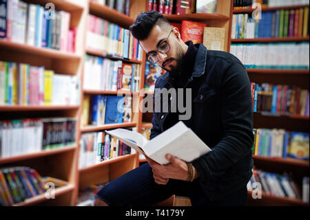 Tall smart étudiant arabe l'homme, porter sur noir veste jeans et lunettes, à Bibliothèque avec livre à mains. Banque D'Images