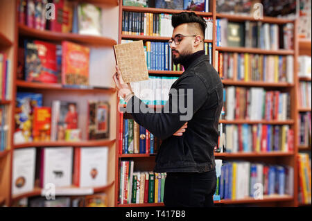 Tall smart étudiant arabe l'homme, porter sur noir veste jeans et lunettes, à Bibliothèque avec livre à mains. Banque D'Images