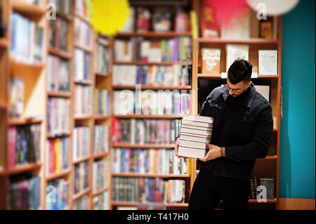 Tall smart étudiant arabe l'homme, porter sur noir veste jeans et lunettes, à Bibliothèque avec pile de livres. Banque D'Images