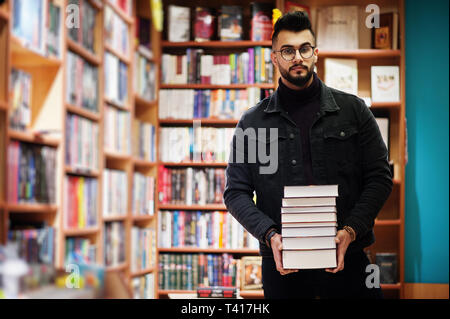 Tall smart étudiant arabe l'homme, porter sur noir veste jeans et lunettes, à Bibliothèque avec pile de livres. Banque D'Images