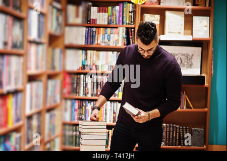 Tall smart étudiant arabe l'homme, de l'usure sur col roulé violet et les lunettes, à Bibliothèque avec pile de livres. Banque D'Images