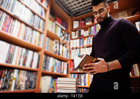 Tall smart étudiant arabe l'homme, de l'usure sur col roulé violet et les lunettes, à Bibliothèque avec pile de livres. Banque D'Images