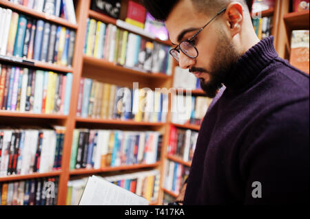 Tall smart étudiant arabe l'homme, de l'usure sur col roulé violet et les lunettes, à Bibliothèque lire livre. Banque D'Images