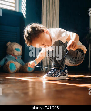 Bébé garçon apprend à s'attacher les lacets de ses baskets en face de la fenêtre. Banque D'Images