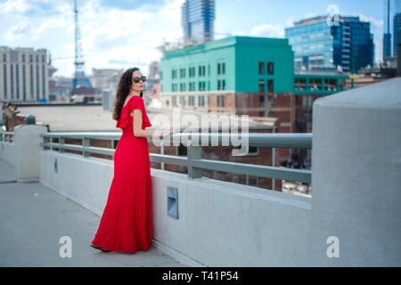 Une belle femme se trouve dans une longue robe rouge contre une ville Banque D'Images